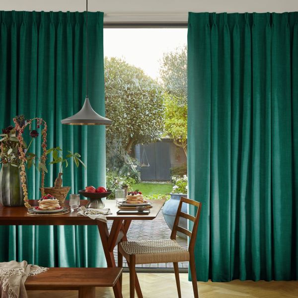 Teal, floor-length pleated curtains hung on a ceiling track, parted centrally to reveal a sunlit garden; a dining table with pancakes and a pendant lamp sits in the foreground.