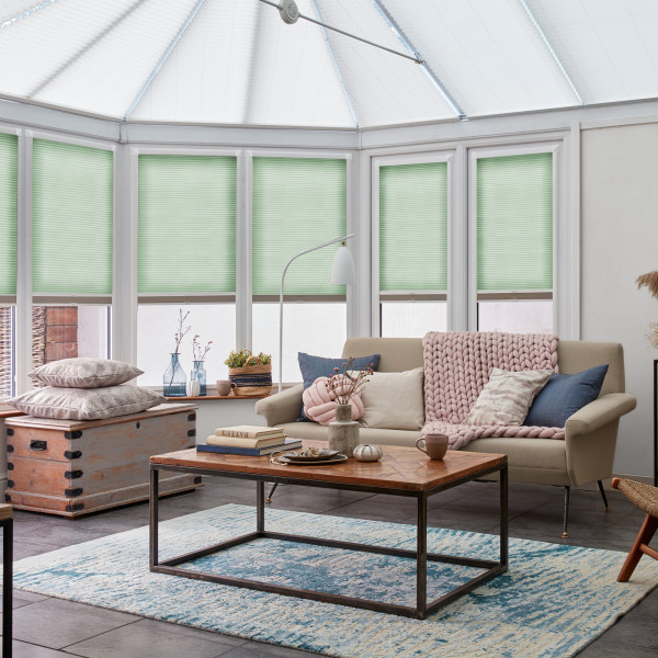 Light-green pleated cellular blinds, lowered halfway across tall white-framed conservatory windows, gently filtering soft daylight; a cozy living area with beige sofa, wooden coffee table, textured rug and plants.