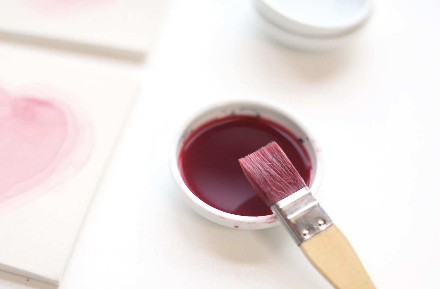 a paint brush with red paint on a white surface next to a handpainted tile.