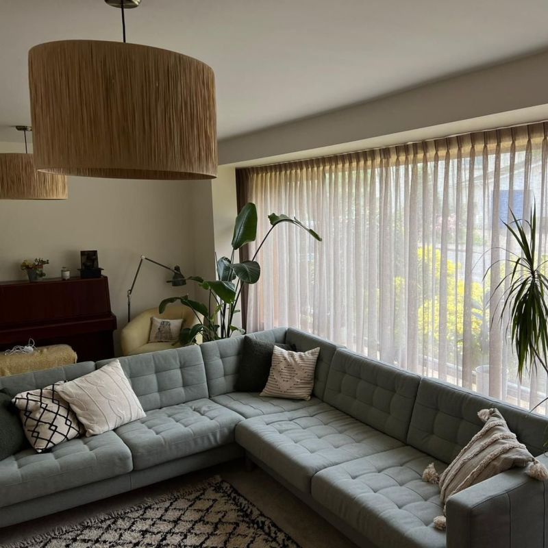 Sheer beige pleated curtains, drawn closed, filtering soft daylight over a corner living room with a gray tufted sectional, tall houseplants, pendant lamps, piano and a garden view beyond.