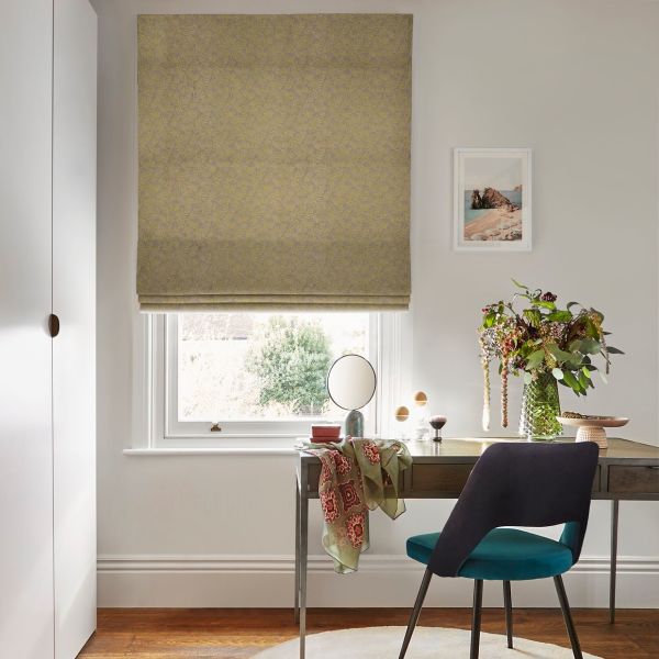 Roman fabric blind (olive‑yellow, leaf pattern), lowered, partially filtering daylight; above a desk with mirror, teal chair and vase of flowers in a neutral, softly lit room.