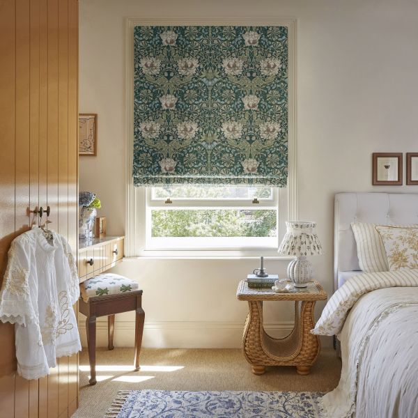 Green floral Roman blind, lowered two-thirds over window, patterned with white blossoms, filtering daylight; bedroom with white upholstered bed, wicker bedside table and wooden wardrobe, leafy outside view.