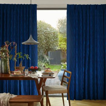 Blue patterned, floor-to-ceiling pleated curtains, parted in the center to reveal a sliding glass door, filtering sunlight; dining table set with pancakes and fruit, pendant lamp, green backyard view.