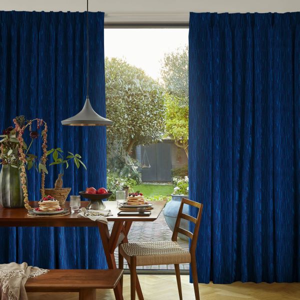 Blue patterned, floor-to-ceiling pleated curtains, parted in the center to reveal a sliding glass door, filtering sunlight; dining table set with pancakes and fruit, pendant lamp, green backyard view.