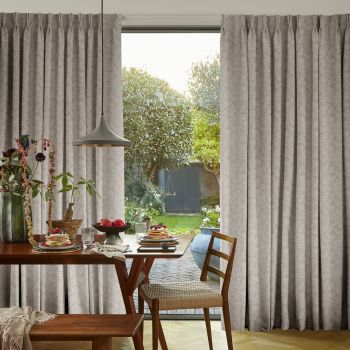 Full-length pleated patterned beige curtains frame a sliding glass door, partially drawn to reveal a sunlit garden; dining table set for breakfast with pancakes, chairs, and a hanging pendant light.