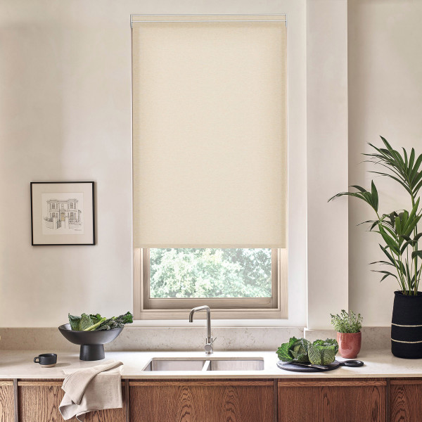 Beige roller blind covers most of a kitchen window above a stainless sink; surrounding countertop features wooden cabinets, a bowl of leafy greens, potted plants, and a framed wall sketch.