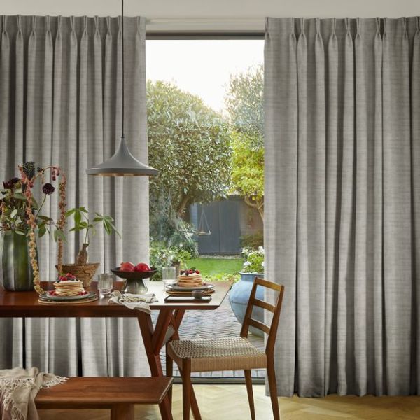 Gray pleated floor-length curtains, hung on a rod and parted centrally to reveal a glass door; filtering daylight over a wooden dining table set for breakfast, a pendant lamp, and a backyard garden.