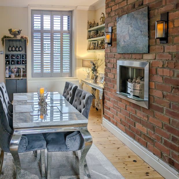 Object â€” white plantation shutters over a tall window with angled louvers; Action â€” mostly closed, filtering daylight; Context â€” dining room with mirrored table, tufted grey chairs, exposed brick wall and recessed fireplace.