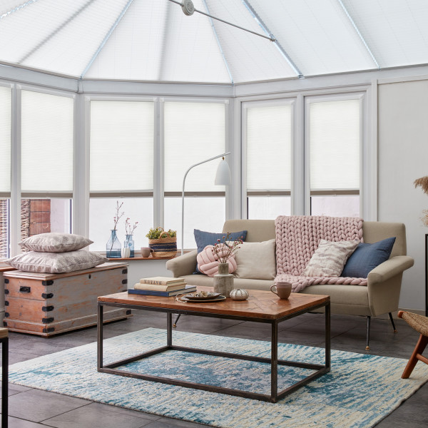 White cellular pleated blinds covering tall bay windows, lowered to mid-height and filtering soft daylight; bright conservatory-style living area with beige sofa, wooden coffee table and blue rug.
