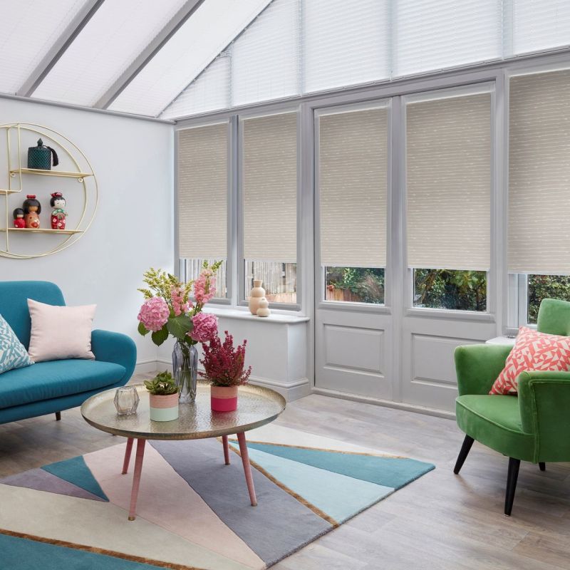 Beige pleated roller blinds covering upper two-thirds of tall conservatory French windows, partially lowered and filtering daylight; sunroom with pastel sofa, green armchair, coffee table, floral arrangements and garden view.