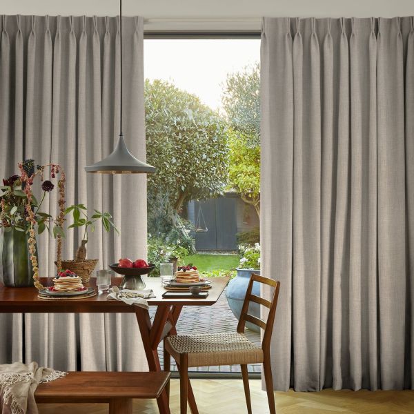 Beige full-length pleated curtains, gathered on each side and slightly parted at center revealing a sunlit garden; inside a dining table set for breakfast with wooden chairs and pendant lamp.
