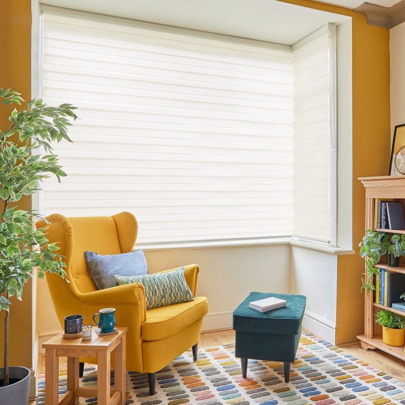 Sheer white roller blinds covering a bay window, fully lowered and diffusing bright daylight; a cozy reading nook with a mustard wing chair, teal ottoman, plant, bookshelf and colorful rug.