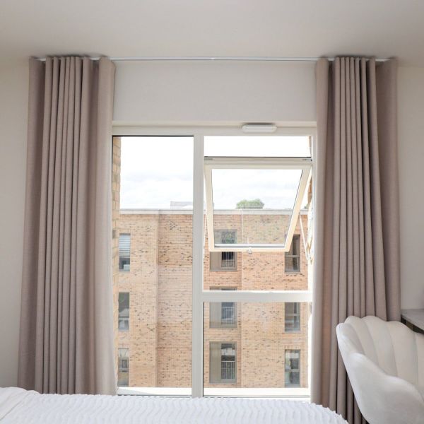 Floor-to-ceiling beige pleated curtains, pulled to each side, framing a white-framed window with its upper casement tilted open; bright bedroom interior, bed foreground, view of brick apartment building.