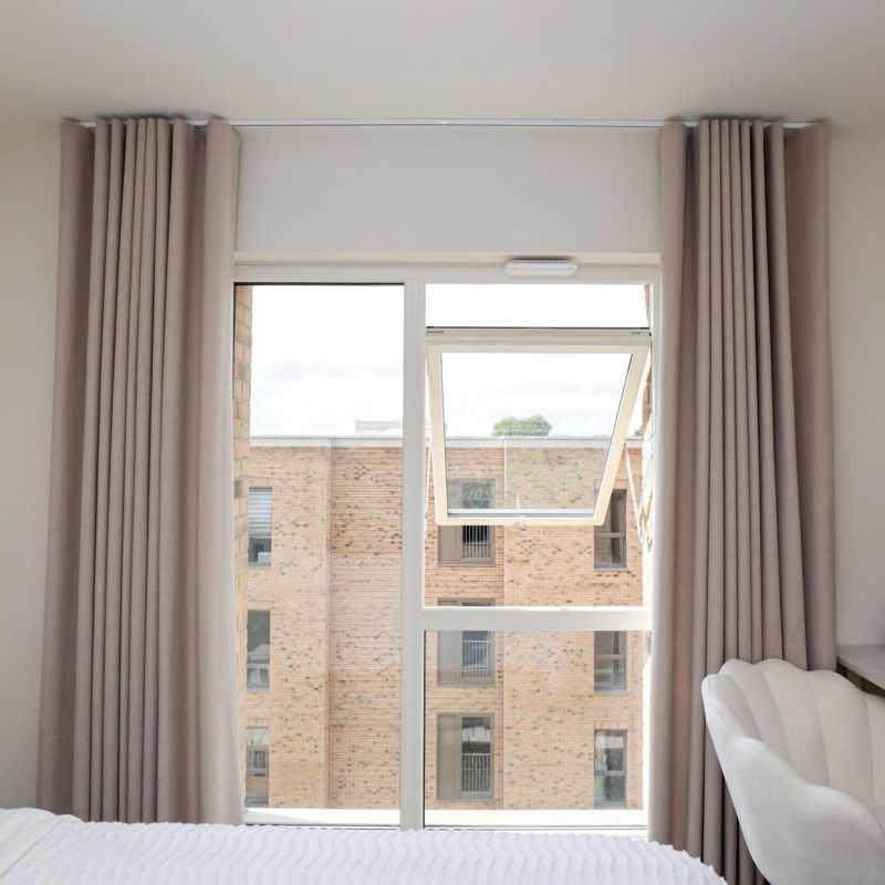 Floor-to-ceiling beige pleated curtains, pulled to each side, framing a white-framed window with its upper casement tilted open; bright bedroom interior, bed foreground, view of brick apartment building.