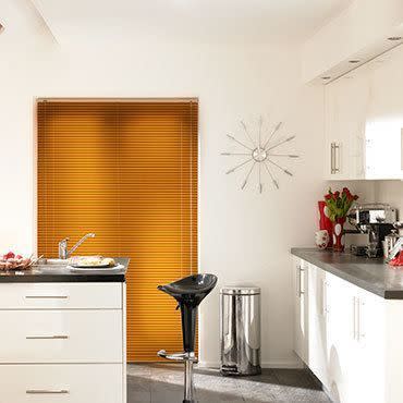 Orange horizontal venetian blinds fully lowered and closed, filtering warm light; modern white kitchen with black countertops, chrome barstool, stainless bin, wall clock, coffee machine and tulips on counter.