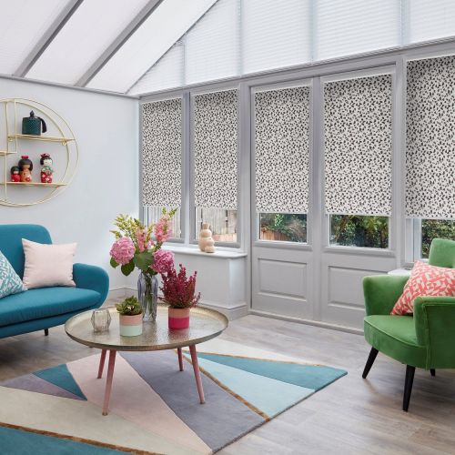 Patterned roller blinds (white with small dark leaf-like speckles) lowered over tall conservatory windows and door, softly filtering daylight; bright living area with teal sofa, green armchair and floral centerpiece.