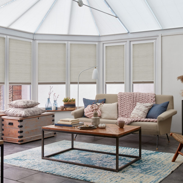 Light beige cellular pleated blinds, lowered across tall conservatory windows and slightly raised at lower edges, filtering soft daylight; cozy sitting room with sofa, chunky pink throw, wooden coffee table.