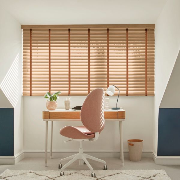 Wooden horizontal Venetian blinds cover a wide dormer window, beige slats with brown vertical tapes, mostly closed and softly filtering sunlight onto minimalist desk in bright attic office.