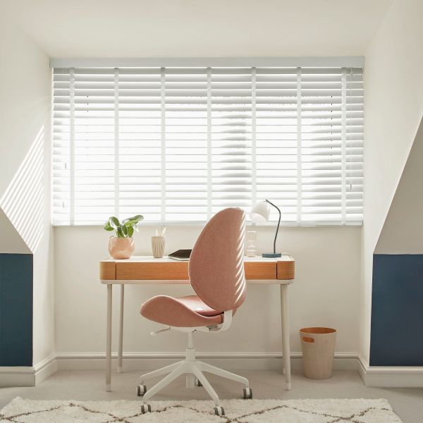 White horizontal Venetian blinds covering a wide window, slats mostly closed and filtering bright daylight; home office with wooden desk, pink swivel chair, lamp, plant, pale walls and blue alcoves.