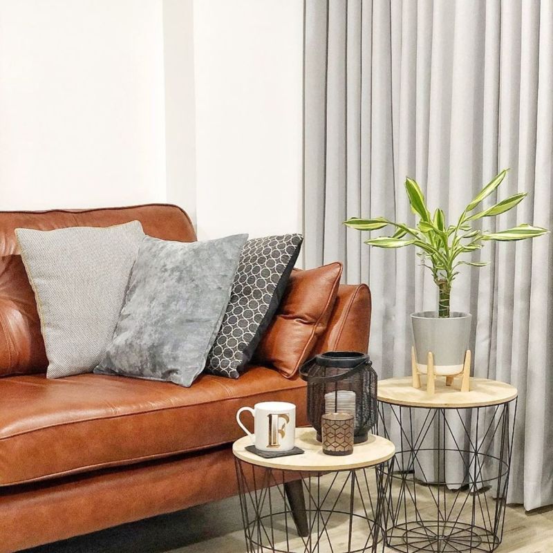 Grey floor-to-ceiling pleated curtains, drawn closed and softly diffusing light; living room with a brown leather sofa, three cushions, wire-frame nesting tables holding a potted plant, lantern, and mug. F