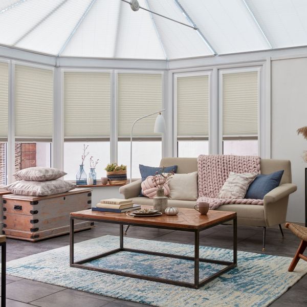 Cream pleated cellular shades, partially lowered across tall bay windows, filtering soft daylight into a bright conservatory-style living room with a light sofa, pink knit throw, wooden coffee table.