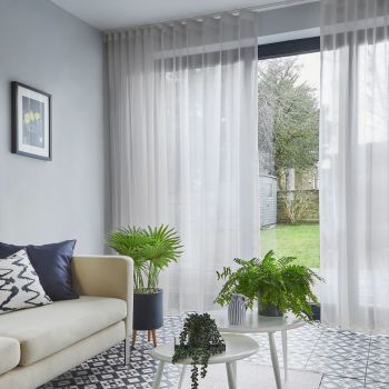 Sheer white, ceiling-mounted floor-to-ceiling curtains with pleats, partially drawn over sliding glass doors, filtering daylight into a living room with pale sofa, patterned tile floor, potted plants and lawn view.