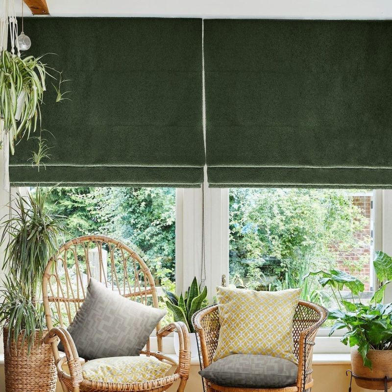 Deep green Roman blinds, lowered and split at the center with a visible pull chain, filtering light; in a sunlit seating nook with wicker chairs, patterned cushions, houseplants and garden view.