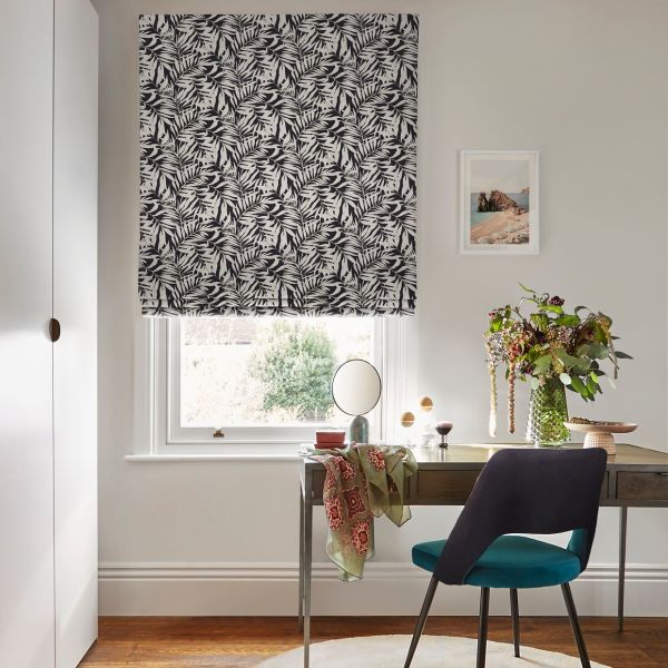 Black-and-white leaf-pattern Roman blind lowered over the window, filtering daylight; a wooden desk with teal chair, small round mirror and vase of foliage beneath, pale walls and hardwood floor.