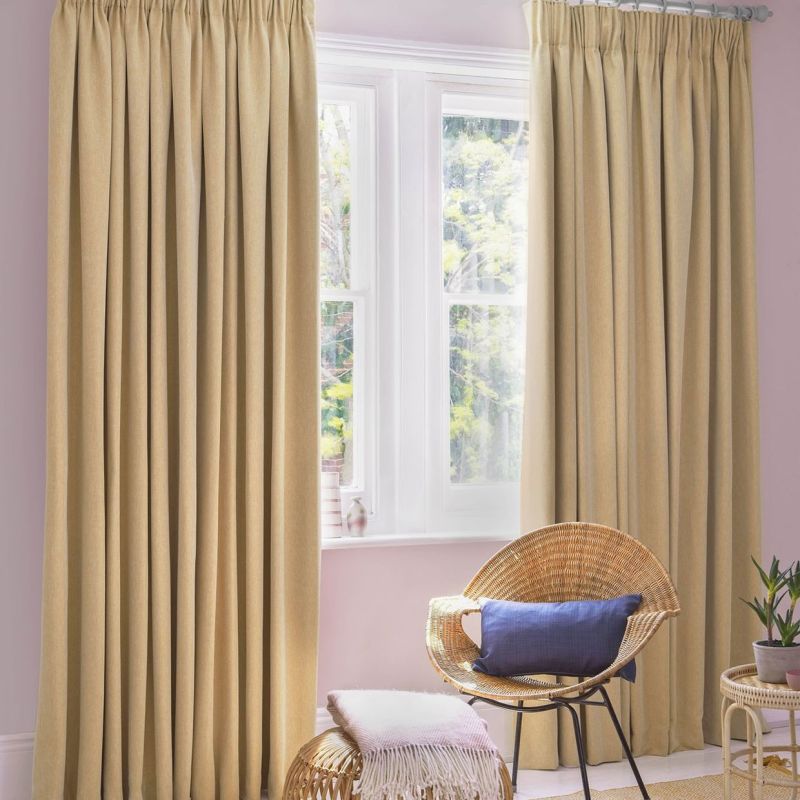 Beige gathered-pleat curtains (full-length, deep vertical folds) â€” mostly drawn, filtering soft daylight through a central white-framed window â€” pastel-pink-walled sitting corner with wicker chair, ottoman, and potted plant.