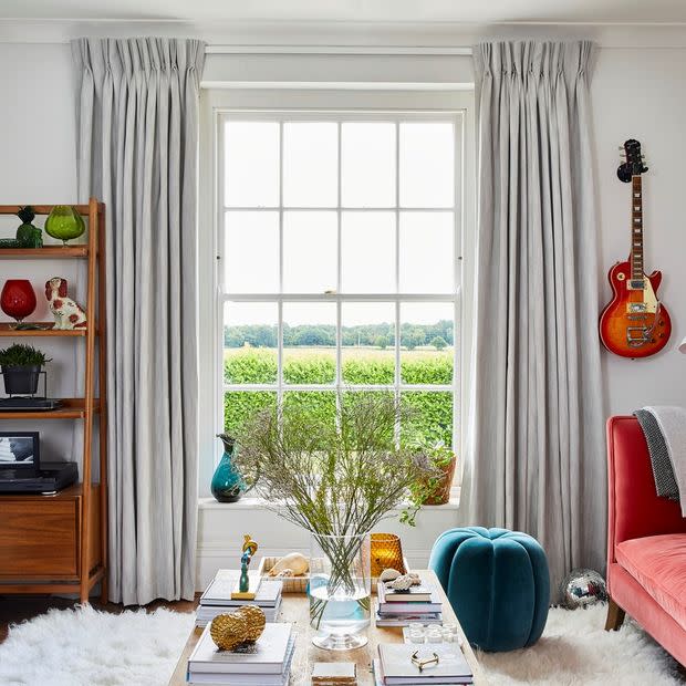 Floor-length pleated grey curtains, hung high and pulled to sides, framing a grid window admitting daylight; cozy living room with coffee table with vase, teal pouf, red sofa and guitar.
