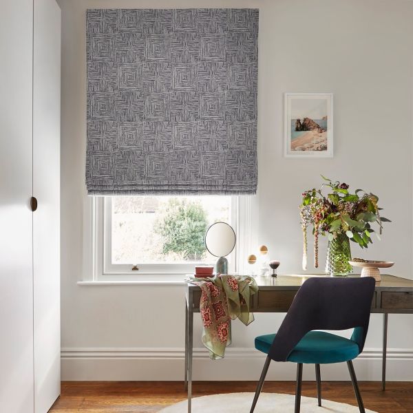 Grey Roman blind with concentric-square pattern lowered over window, filtering daylight; above wooden desk with mirror, brushes, vase of flowers and teal-upholstered chair in neutral home office.