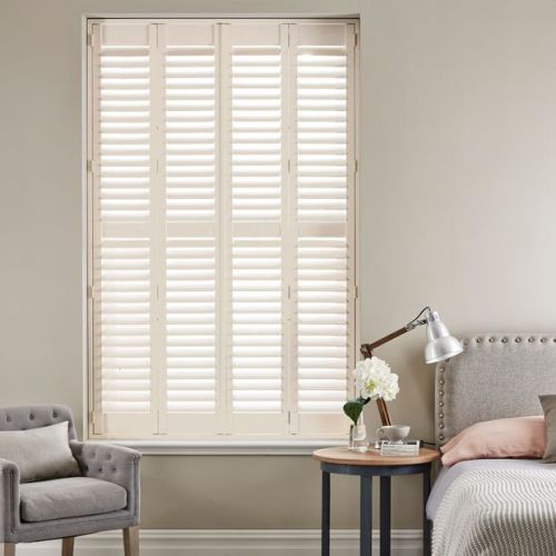 White plantation shutters covering a tall window, louvered panels closed and filtering soft daylight; set in a neutral-toned bedroom with upholstered bed, bedside table, lamp, and armchair.