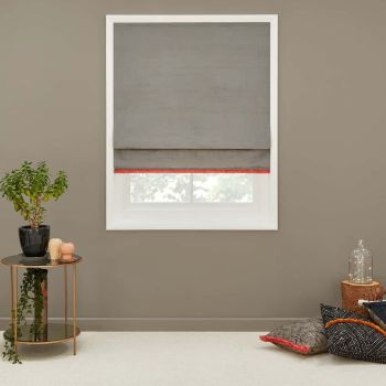 Gray fabric Roman shade with thin red trim lowered over a white-framed window, filtering soft daylight; taupe living-room wall, side table with potted plant and floor cushions nearby.