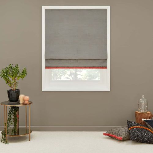 Gray fabric Roman shade with thin red trim lowered over a white-framed window, filtering soft daylight; taupe living-room wall, side table with potted plant and floor cushions nearby.