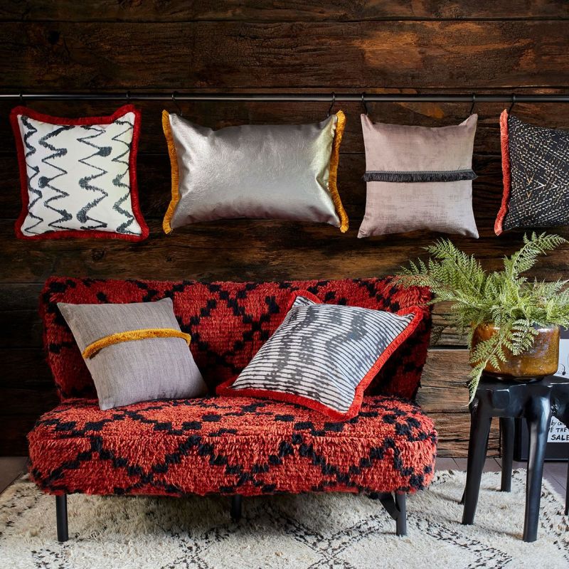 Decorative throw pillowsâ€”fringed, metallic, and patternedâ€”hang from a horizontal rod and rest on a low red-and-black shag loveseat; rustic wood wall, cream patterned rug, black stool with potted fern.