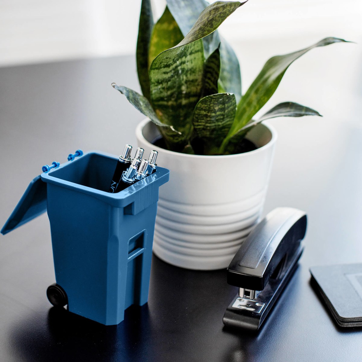 mini blue recycling cart on a desk with pens inside sitting beside a plant and stapler