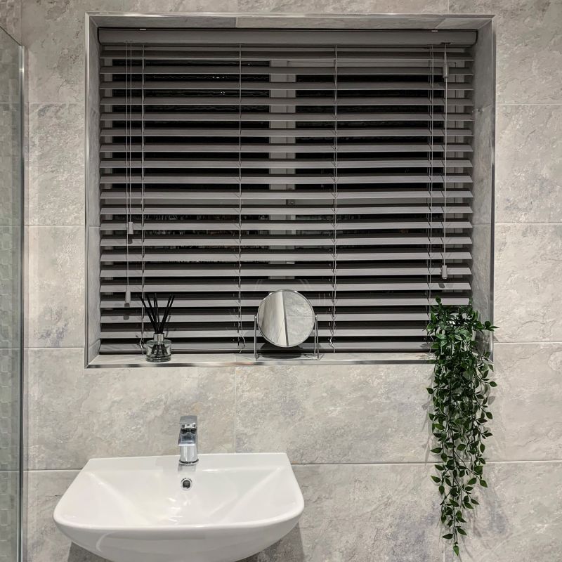 Grey horizontal venetian blinds, fully closed with visible cords and slats, filtering dim light through a recessed tiled bathroom window; white sink, small round mirror, diffuser, trailing plant on sill.