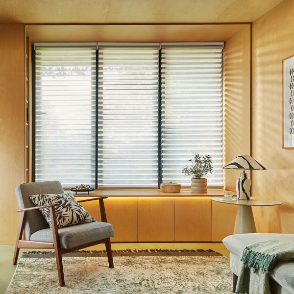 Layered horizontal zebra blinds covering a three-panel window, lowered and filtering pale daylight; they cast soft stripes across a warm wood-paneled living nook with armchair, side table, and plant.