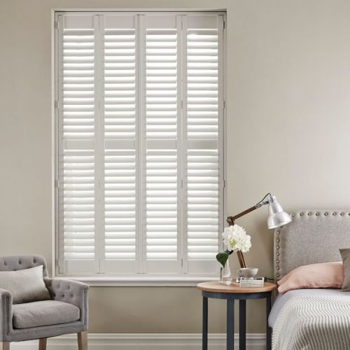 White plantation shutters covering a tall windowâ€”four framed panels with horizontal slats, closed and filtering soft daylightâ€”set in a neutral bedroom corner with upholstered headboard, bedside lamp, vase, and armchair.