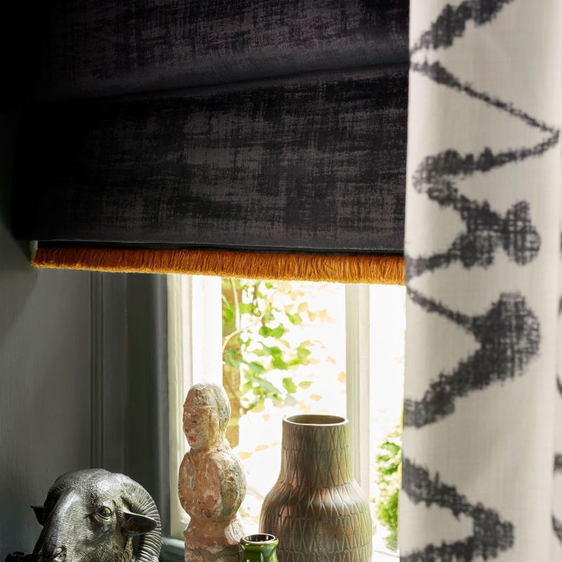 Black Roman blind with orange fringe lowered over a window; patterned curtain at right. Soft daylight filters onto a sill of vases and sculptures, with leafy exterior visible.