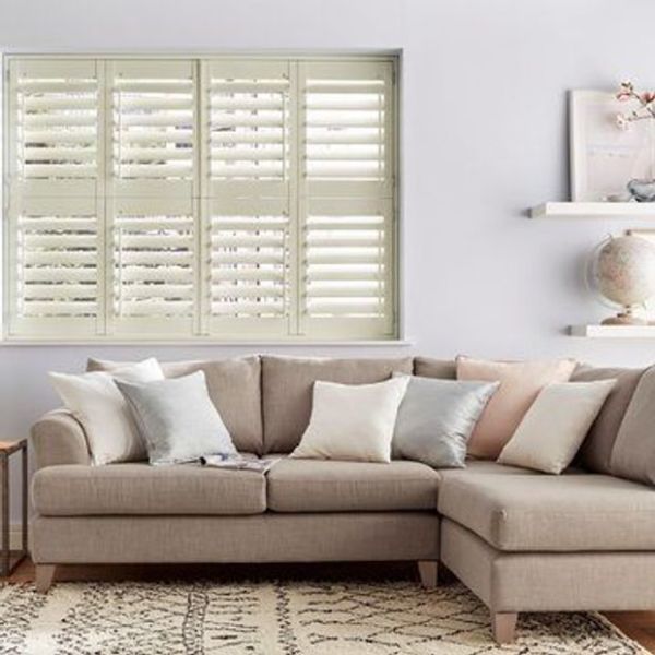 White plantation shutters covering a wide window, louvered slats tilted to filter soft daylight; set above a beige L-shaped sectional with pastel cushions in a bright, neutral living room.