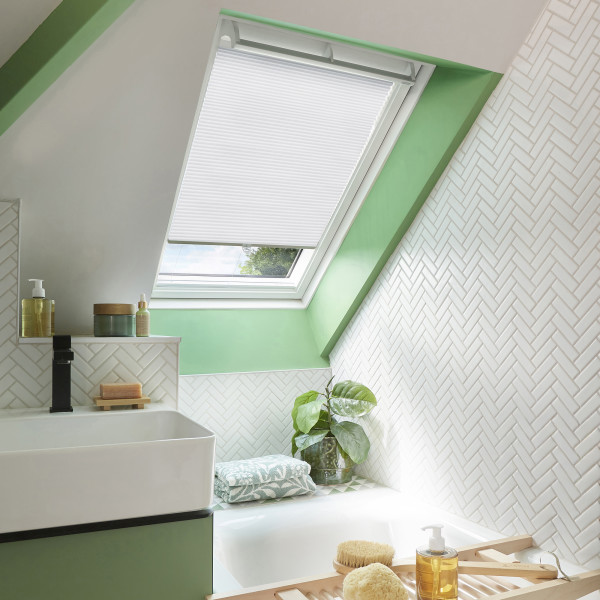 A white pleated skylight blind, fitted to an angled roof window and lowered about three-quarters, filters daylight; bathroom with mint-green trim, white herringbone tiles, sink, bathtub, plant and toiletries.