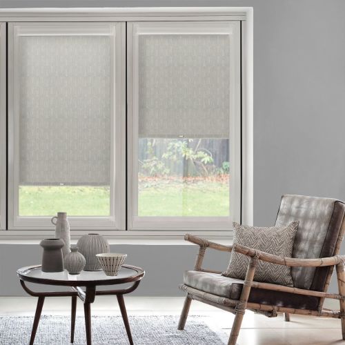 Two patterned roller blinds, lowered to cover most of a double window with translucent lower sections, filtering daylight; gray living room with rattan armchair, side table, decorative ceramics, and garden view.