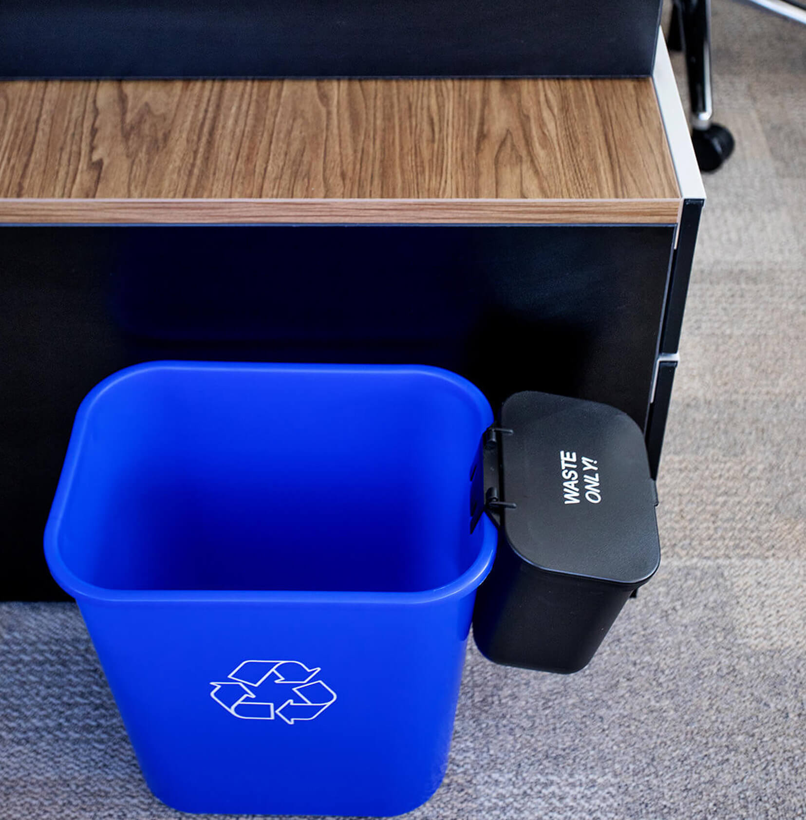 blue plastic recycling bin with a white mobius loop printed on the sides including a hanging waste container with hinged lid sitting beside an office desk