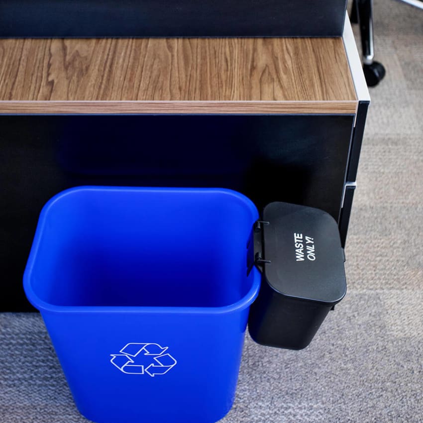 blue plastic recycling bin with a white mobius loop printed on the sides including a hanging waste container with hinged lid sitting beside an office desk