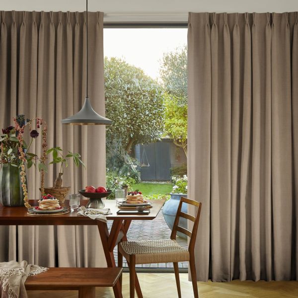 Beige pleated floor-length curtains, hung on a track and parted centrally, framing a sunlit backyard; wooden dining table set for breakfast with a pendant lamp in the foreground.