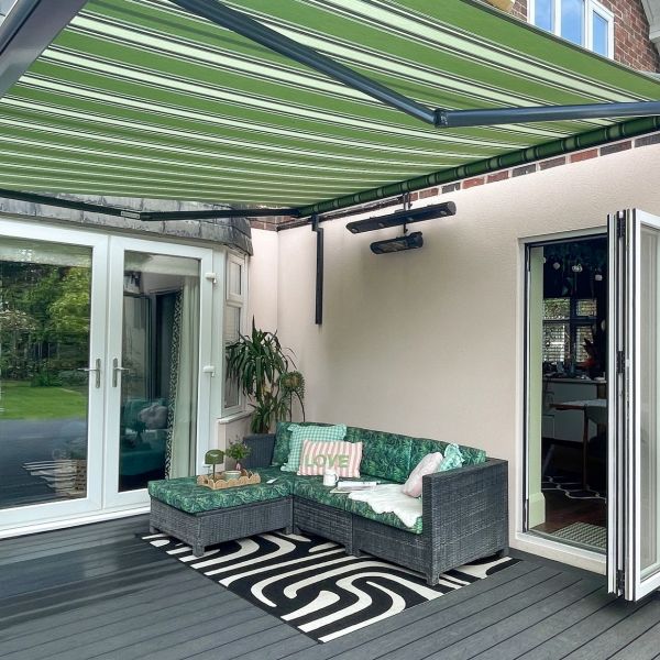 Green-and-white striped retractable awning overhead, fully extended and shading a patio; rattan L-shaped sofa with tropical cushions beside open folding doors on gray decking and a potted plant.
LOVE