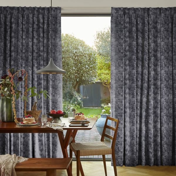 Dark textured pleated curtains, drawn aside centrally, framing an open glass door; daylight and a green garden visible beyond; foreground dining table set for breakfast beneath a pendant lamp.