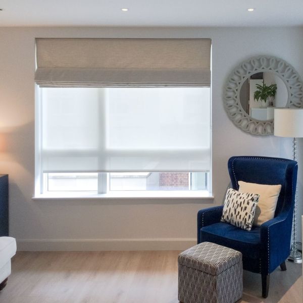 Beige fabric roman shade with a lowered white roller blind, both covering a large rectangular window; filtering soft daylight in a living-room corner with a blue armchair, ottoman, mirror.