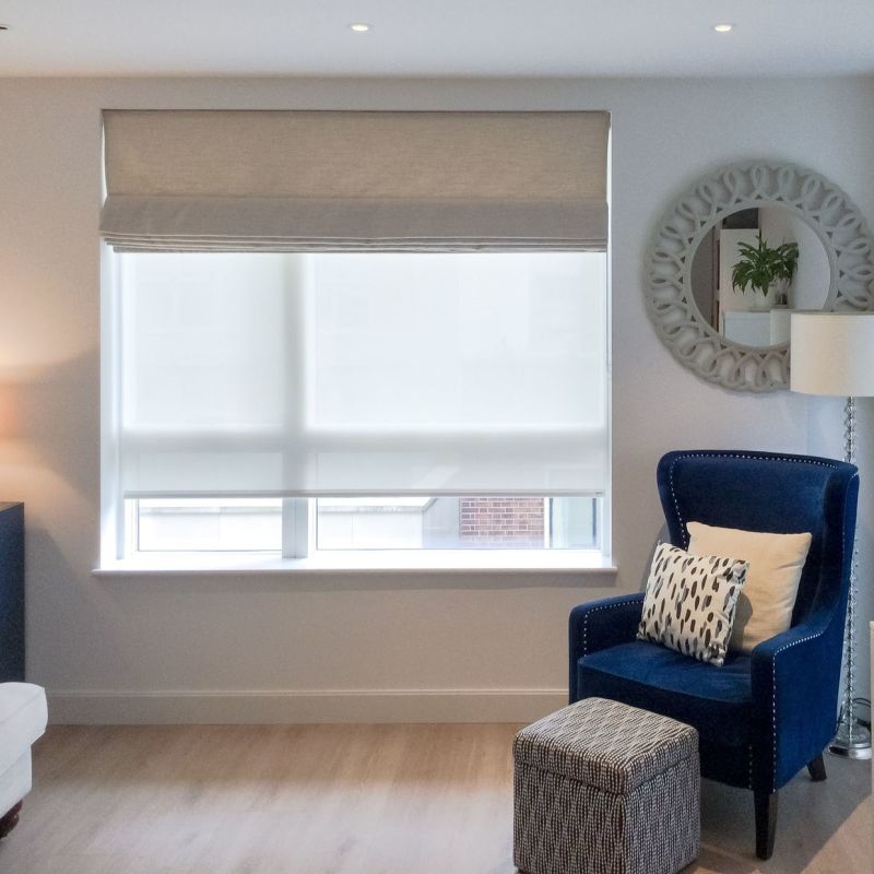 Beige fabric roman shade with a lowered white roller blind, both covering a large rectangular window; filtering soft daylight in a living-room corner with a blue armchair, ottoman, mirror.